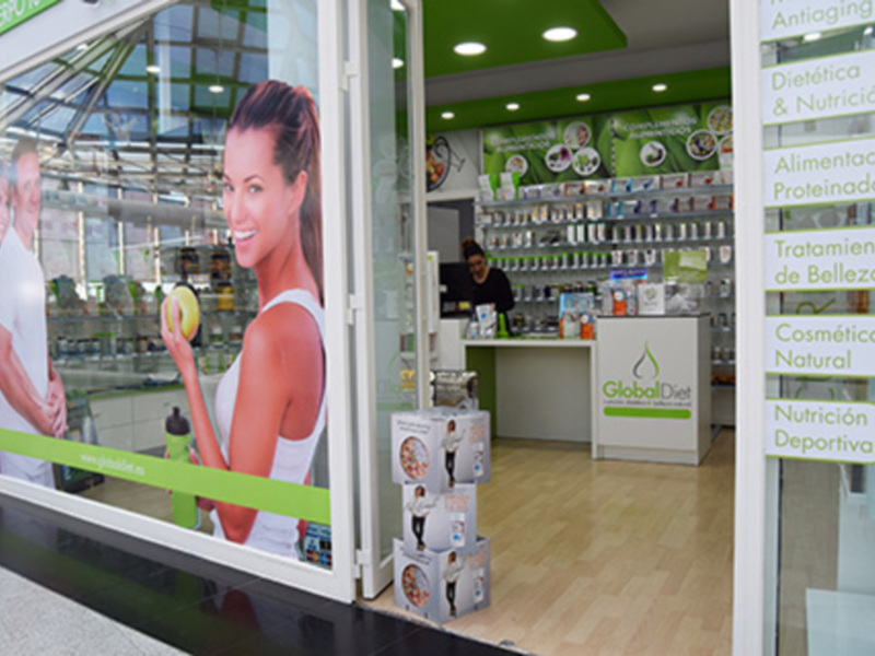 Tienda de belleza con productos de cosmética natural y deportiva. Se puede ver una mujer sonriente con una botella de agua en la mano, y el interior de la tienda con estanterías llenas de productos.