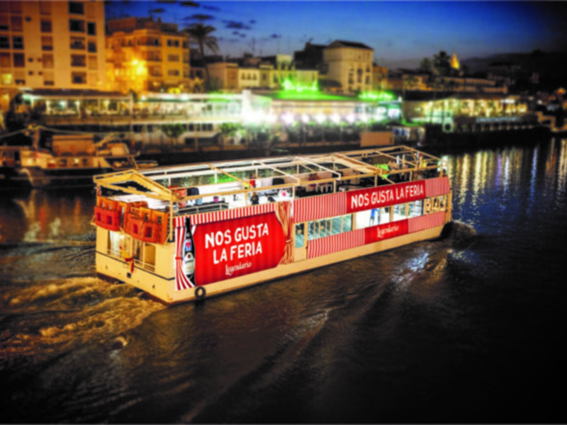 Barco flotando en el río con iluminación nocturna, mostrando publicidad de "Nos Gusta La Feria".