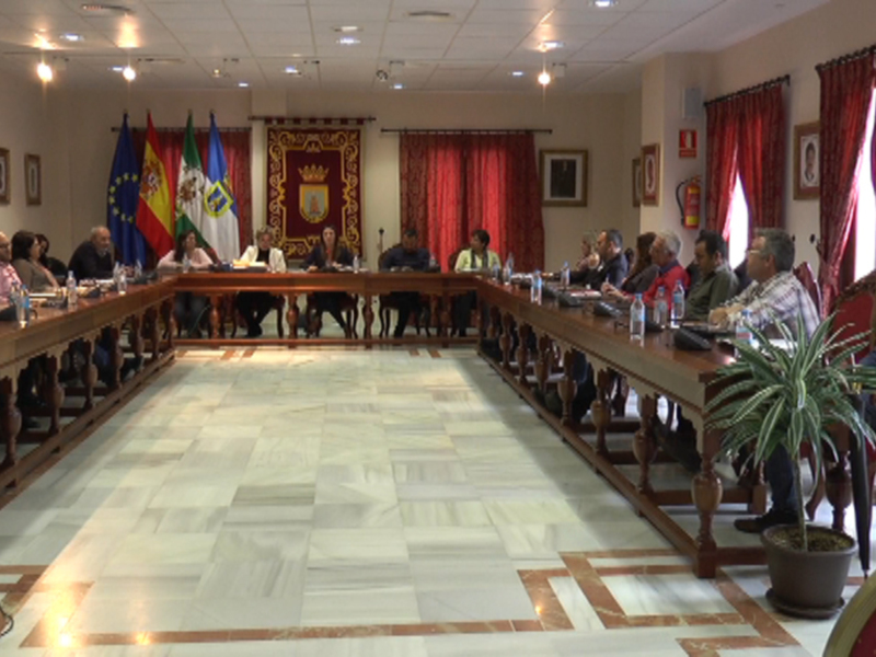 Sala de reuniones con una larga mesa central, banderas de España y Europa, y personas sentadas en sillas rojas. El suelo es de mármol y hay dos plantas en macetas en la parte frontal de la sala.