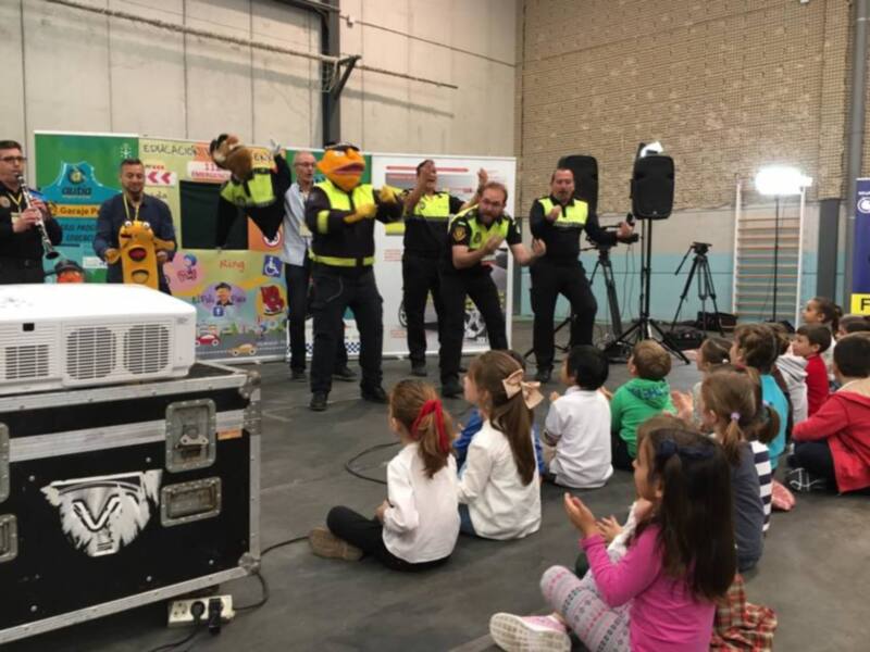Niños escuchando a una mascota y a policías en una actividad educativa en una escuela. El ambiente es animado y los niños están interesados en lo que están aprendiendo.