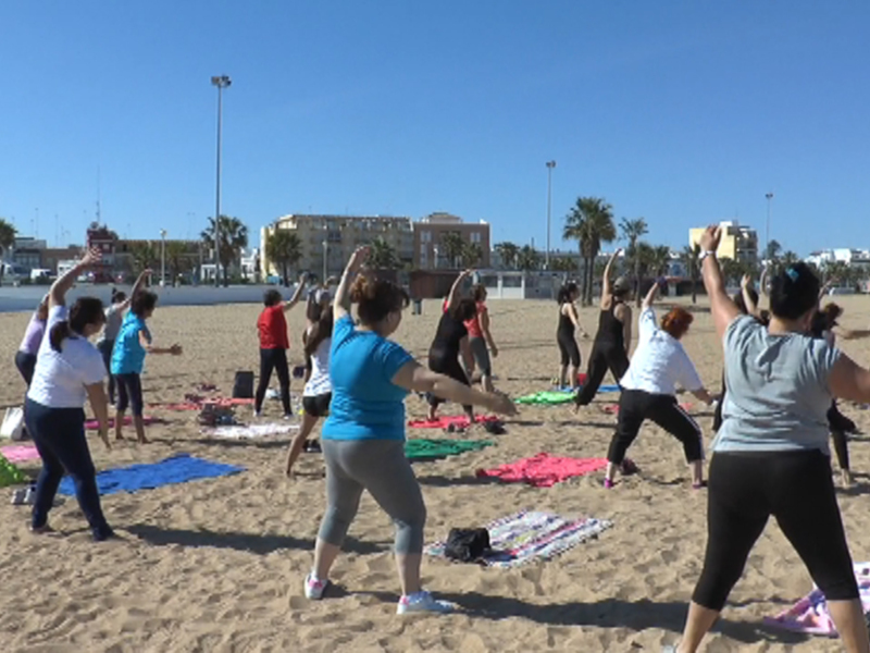 Clase de yoga en la playa con personas realizando ejercicios de estiramiento bajo el sol.