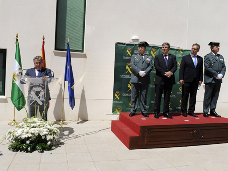 Una ceremonia formal con tres hombres en uniforme y trajes, junto a una bandera y un escudo de armas, en un entorno formal con un escudo de armas en el fondo.