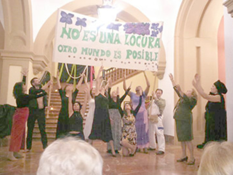 Una escena teatral con un grupo de actores levantando un cartel que dice 'NO ES UNA LOCURA OTRO MUNDO ES POSIBLE' en un interior con arcos y columnas.