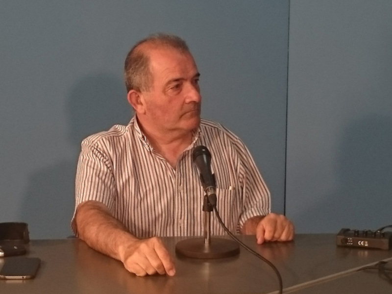 Hombre sentado en una mesa con un micrófono, posiblemente en una conferencia o entrevista.