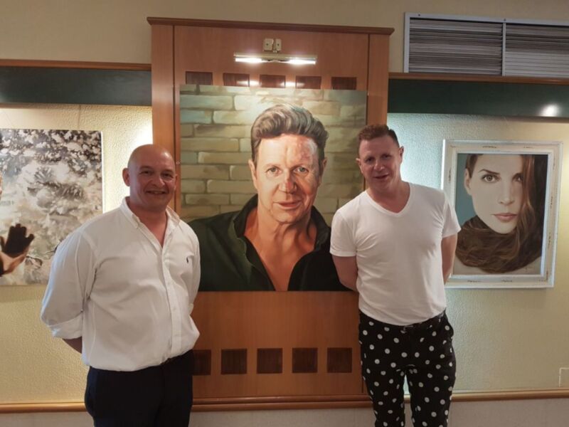 Dos hombres posan junto a una pintura de una persona en una sala de exposiciones.