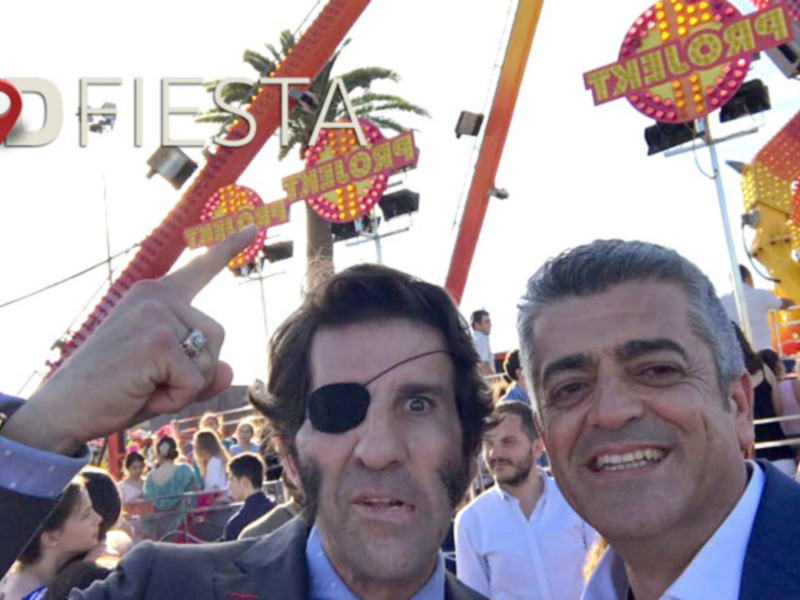 Dos hombres se toman una selfie en una feria con atracciones de fondo y el logo 'ADFIESTA' en la parte superior.