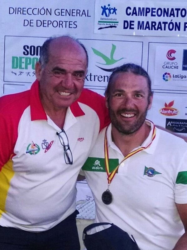 Dos personas sonrientes en un evento deportivo, posando frente a un cartel con logos de patrocinadores y el nombre del evento.