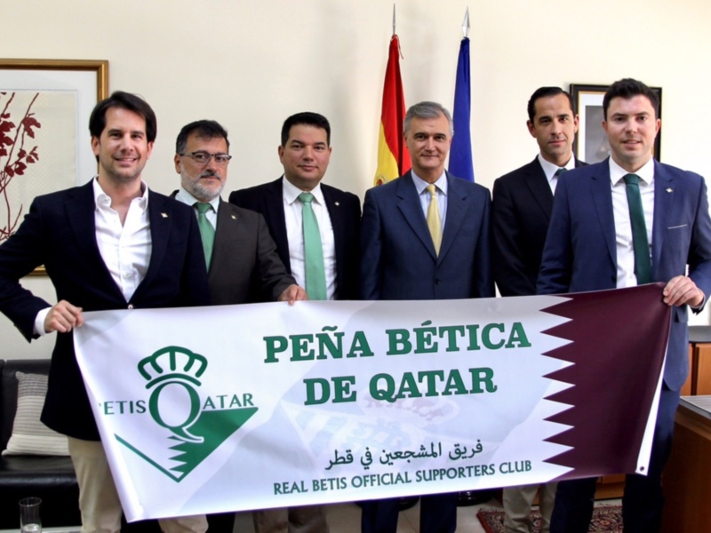 Cuatro hombres en trajes sostienen una bandera de la Peña Betis Bética de Qatar en una sala formal con banderas de España y Qatar en el fondo. El texto en la bandera es 'Peña Betis Bética de Qatar' en español y árabe, junto con el logotipo de Real Betis.