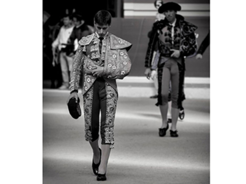 A matador in traditional bullfighting attire, including a patterned jacket and a sombrero, walks on a bullring floor.