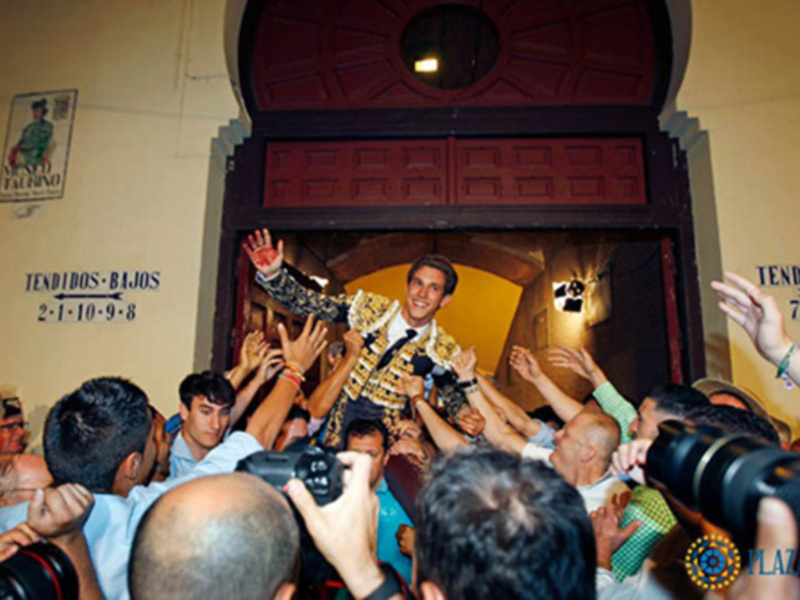 Un hombre en traje de corrida es portado por una multitud en una celebración festiva, con una puerta grande y decorada en el fondo.