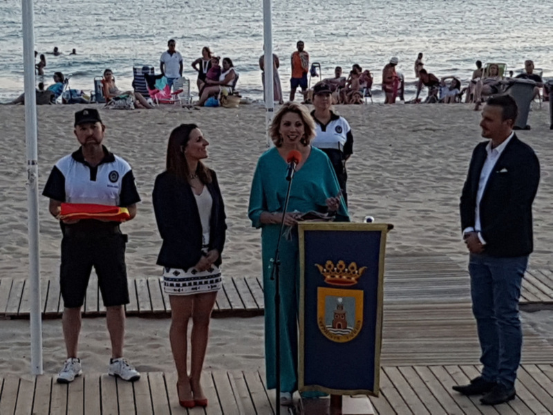 Una mujer habla en un podio con un escudo de armas, mientras dos personas la observan en una playa con personas disfrutando del mar al fondo.