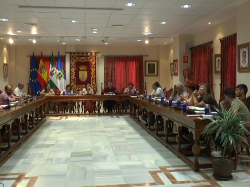 Reunión de consejo municipal en una sala formal con banderas y escudos oficiales.