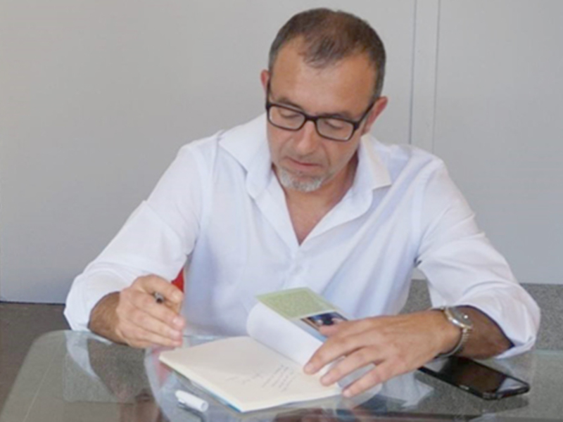 Un hombre en una camisa blanca está sentado en una mesa de cristal, escribiendo en un libro abierto. A su lado, hay un teléfono móvil. El fondo es una pared blanca.