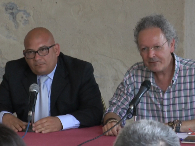 Dos hombres sentados detrás de un micrófono en una mesa, participando en una reunión o conferencia.