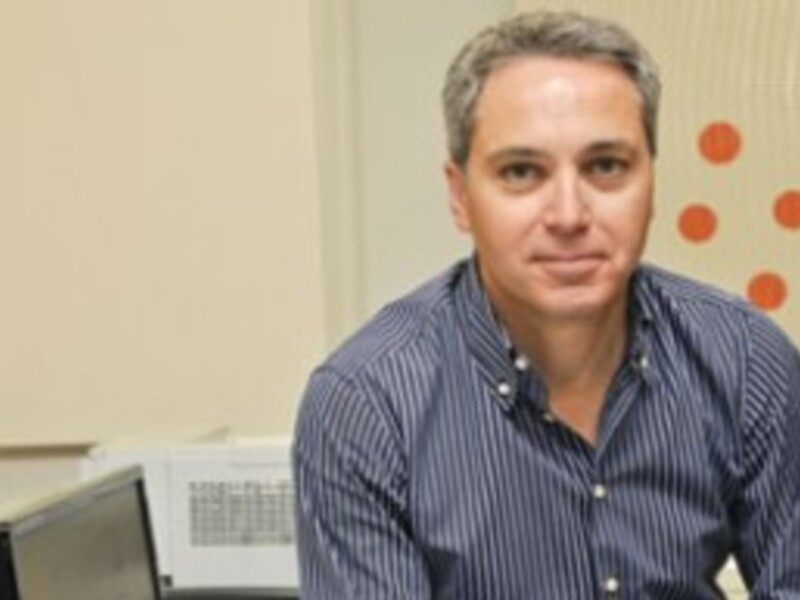Un hombre con cabello gris corto, mirando directamente a la cámara, con una camisa azul a rayas y una pared de fondo con un panel de ventilación y una pared amarilla con puntos rojos.