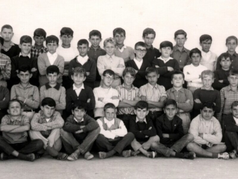 Grupo de niños en una foto de grupo de escuela, con uniformes y camisas de diferentes colores. Algunos niños llevan gafas y otros tienen el brazo cruzado. El fondo es blanco y hay dos adultos en la parte superior de la foto.