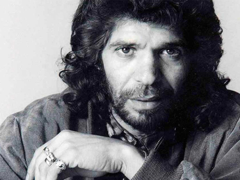 A black and white portrait of a man with long hair and a beard, wearing a jacket and a ring on his finger.