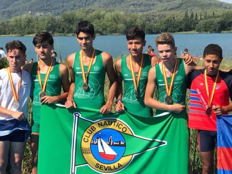 Equipo de remo del Club Náutico Sevilla celebrando con medallas y la bandera del club en un entorno acuático.