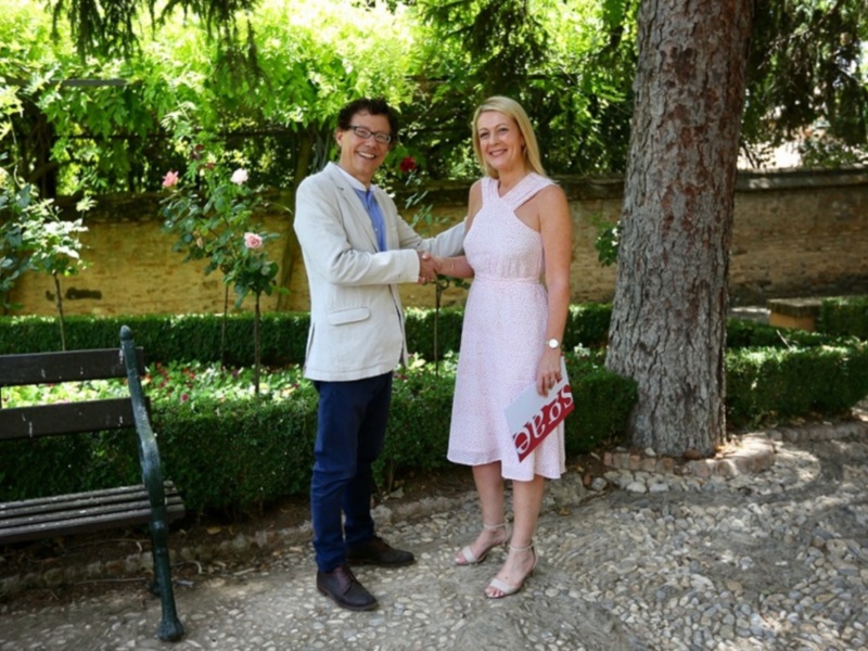 Dos personas saludándose en un jardín soleado rodeado de flores y árboles.