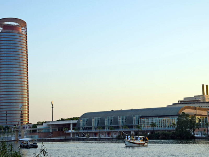 Un barco pequeño navegando cerca de una torre de oficinas moderna en un entorno urbano con edificios y árboles al fondo, durante un día soleado.