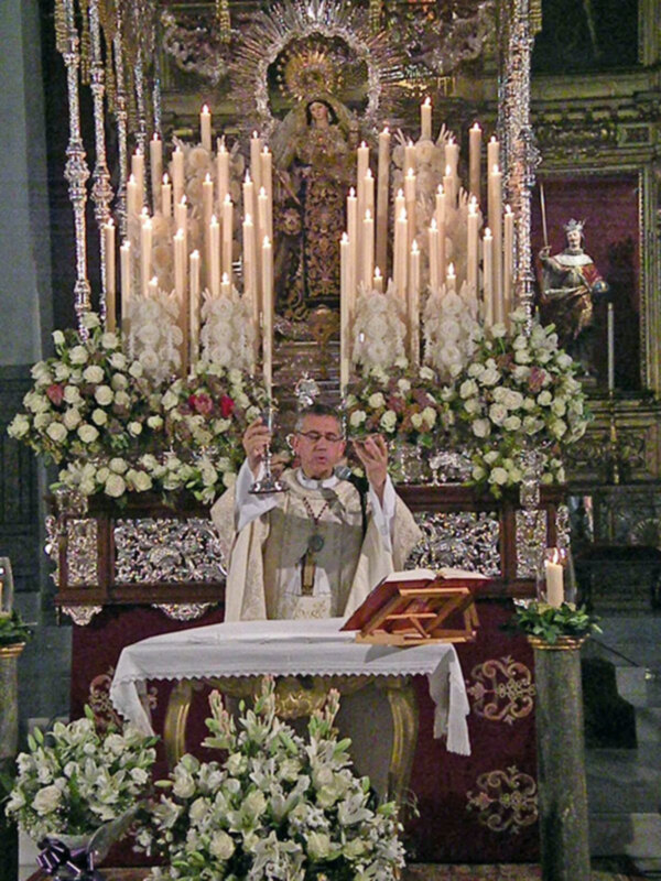 Sacerdote oficiando una misa en un altar adornado con velas y flores, en un entorno religioso.
