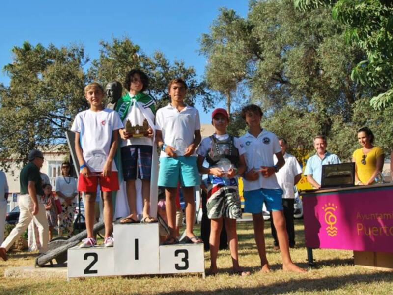 Tres jóvenes ganadores de un torneo deportivo posan en el podio, con trofeos en sus manos, mientras otros participantes y espectadores observan en el fondo.