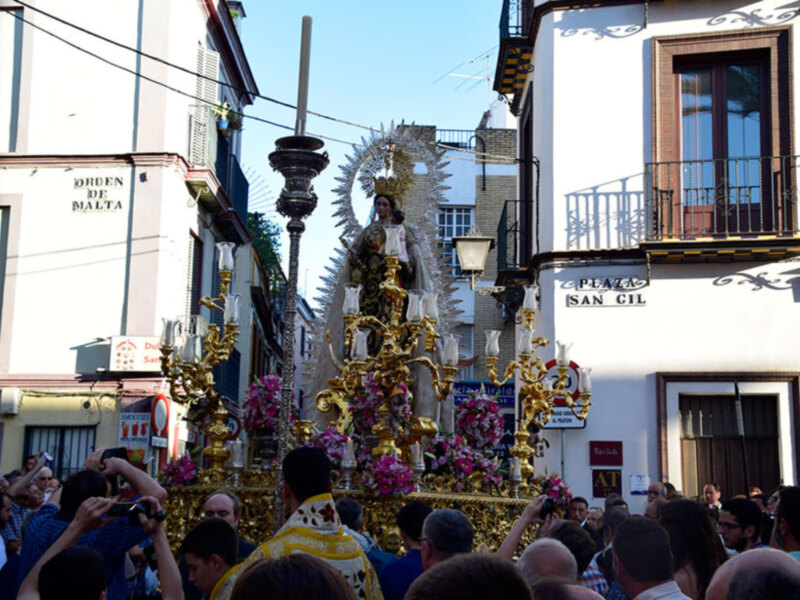 Fotografía de una procesión religiosa con una imagen de la Virgen en el centro, rodeada de flores y luces, mientras una multitud de personas la observa desde la calle.