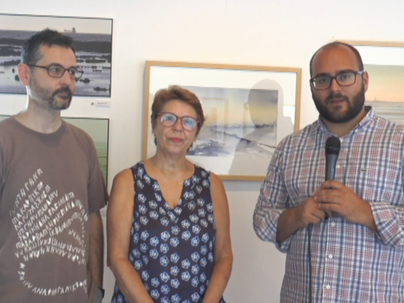 Tres personas en una presentación, con un hombre sosteniendo un micrófono y dos personas de pie a su lado. En el fondo, hay dos cuadros en la pared.
