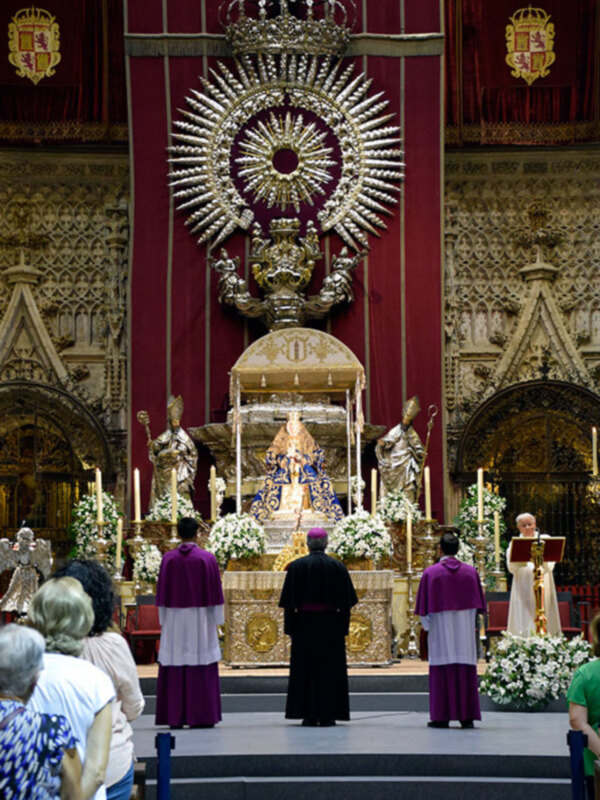 Elegante ceremonia religiosa con un altar dorado y una corona en el centro, rodeada por personas vestidas con vestidos de color morado y negro, con flores blancas y velas en la parte frontal.