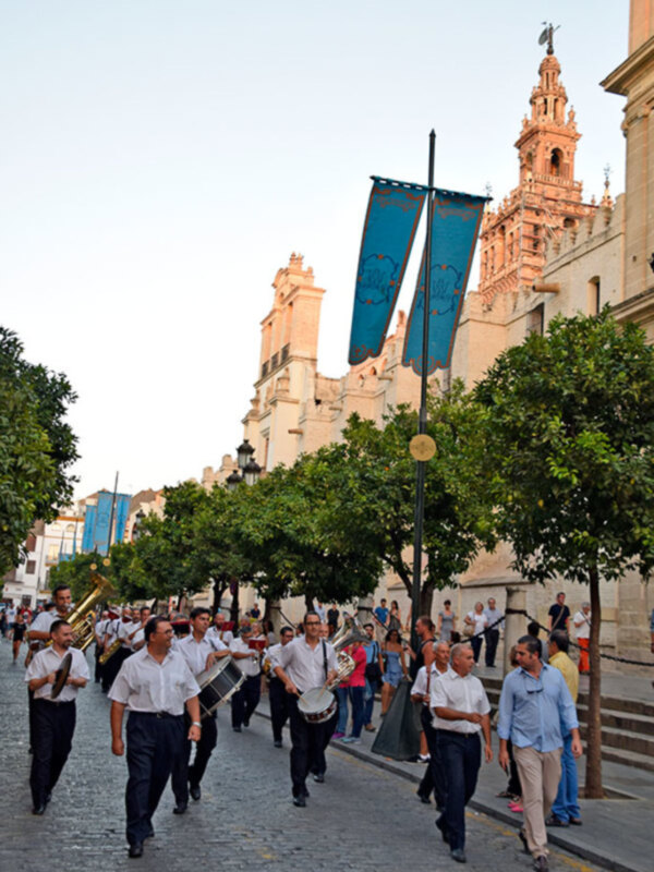Desfile de músicos en una calle de la ciudad, con banderas azules y un edificio histórico al fondo.