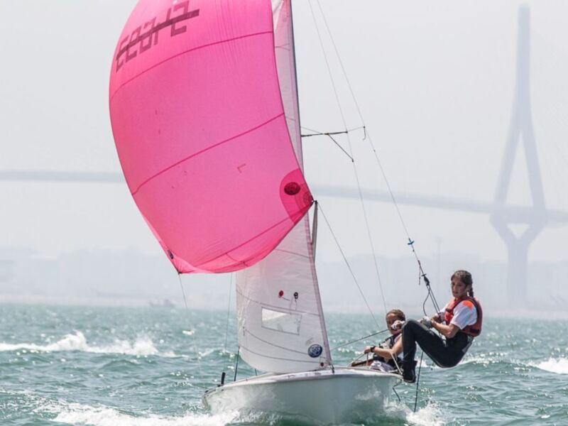Dos regatistas disfrutan de una carrera en velero con vela rosa vibrante en el mar, con un puente en el fondo.