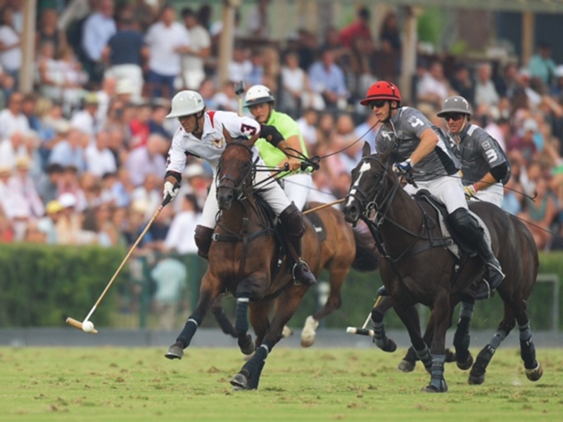 Jugadores de polo en acción durante un partido, con caballos y palos en mano, en un escenario con espectadores en el fondo.