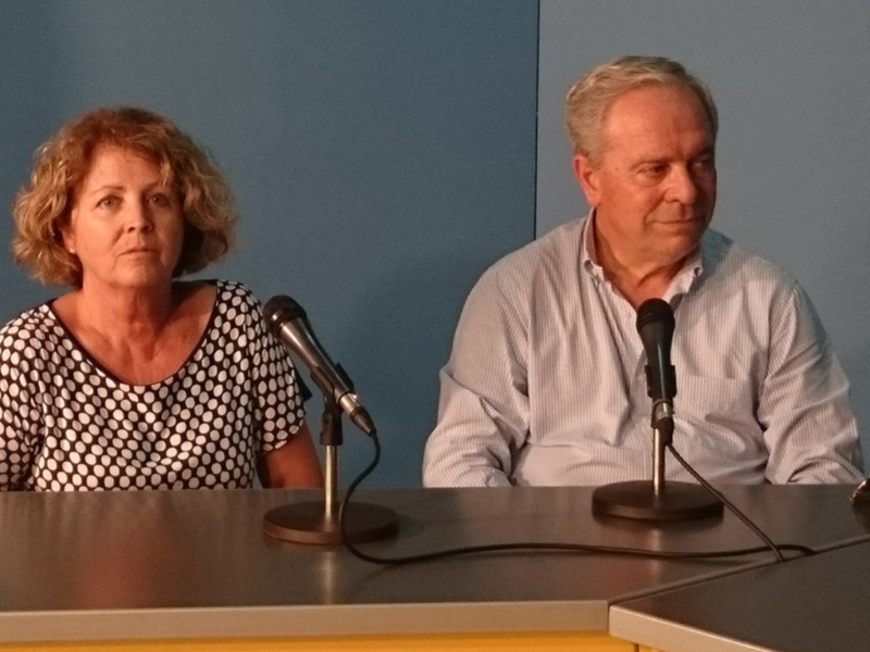 Persona y hombre sentados en una mesa con micrófonos, posiblemente en una conferencia o entrevista.