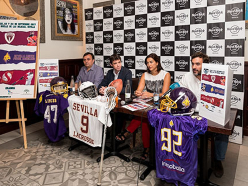Un evento de presentación de equipos de fútbol americano y lacrosse con camisetas y cascos en una mesa con un fondo de logotipos de Hard Rock.