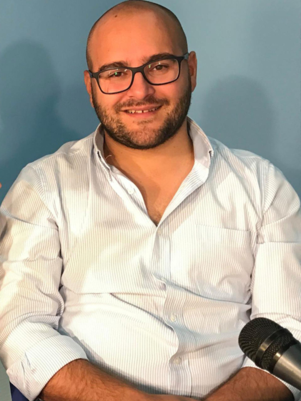 Un hombre con barba y gafas sonríe mientras sostiene un micrófono.