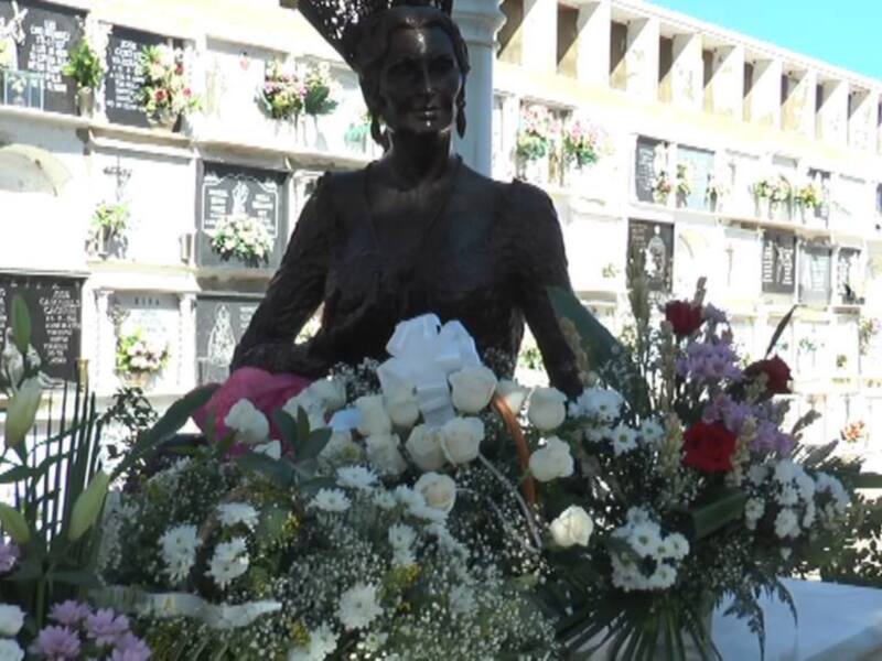 Estatua de una mujer en un cementerio, rodeada de flores y enterradas en nichos con nombres en las paredes detrás.