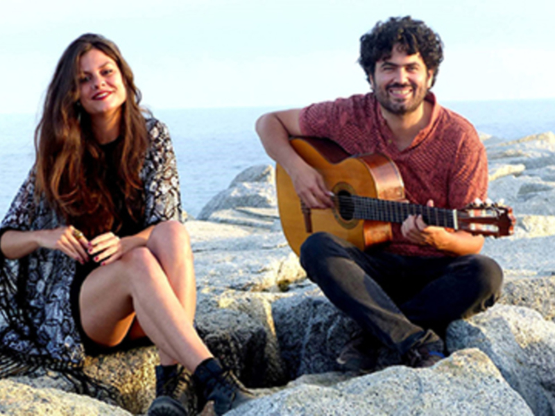 Dos músicos disfrutan de un momento tranquilo junto al mar, con una guitarra y una sonrisa.