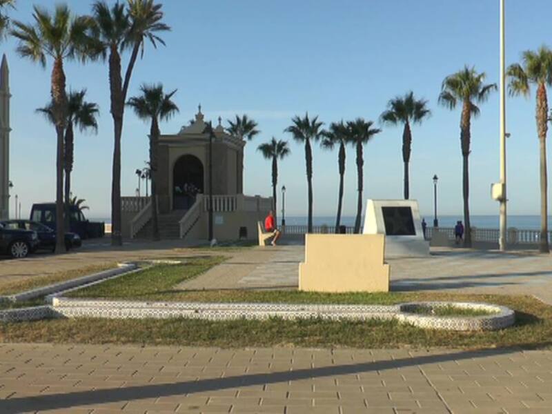 Patio con palmeras y un monumento en el centro, con una vista del mar en el fondo. La imagen muestra un ambiente relajado y soleado, con una mezcla de naturaleza y arquitectura.