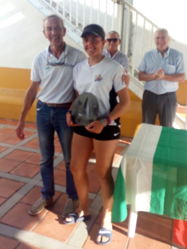 Una mujer en ropa deportiva posa con un casco, mientras un hombre la observa sonriendo. Detrás de ellos, dos hombres más sonríen y aplauden. En el fondo, se puede ver una bandera verde y blanca.