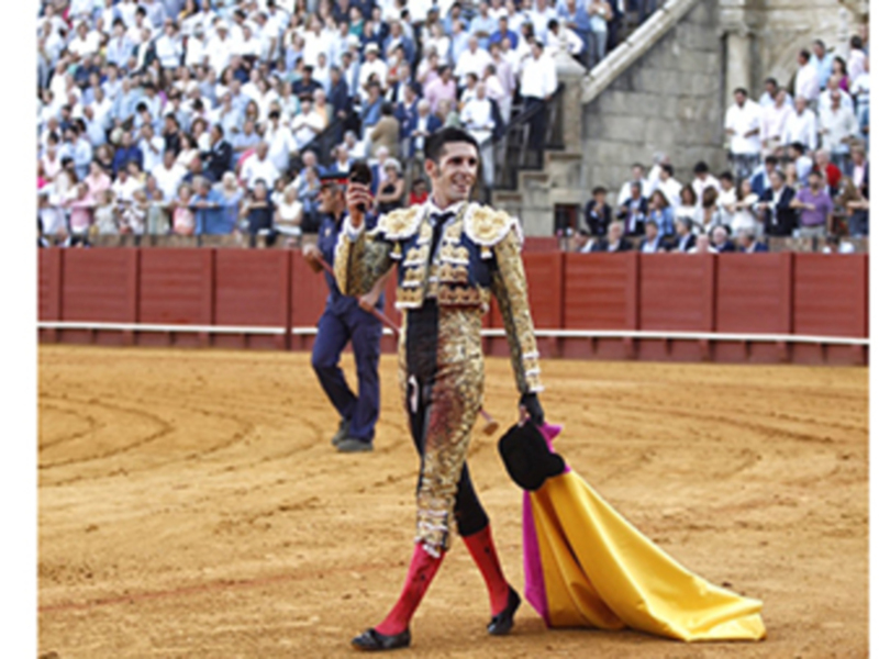 Matador in traditional Spanish bullfighting attire performing with a red and yellow cape in a sandy arena, with a crowd of spectators in the background.