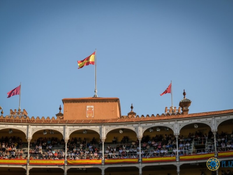 Plaza de Toros de Ronda con bandera española y público en la grada bajo un cielo azul claro.