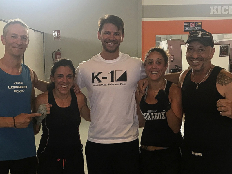 Grupo de personas en una escuela de boxeo, posando con una camiseta K-1 en el centro. La escuela tiene una pared con carteles de boxeo y un reloj en la pared.