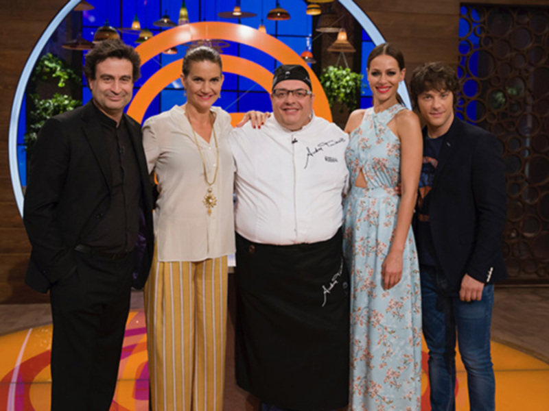 Grupo de personas en un estudio de televisión con un fondo decorativo y un chef en el centro.