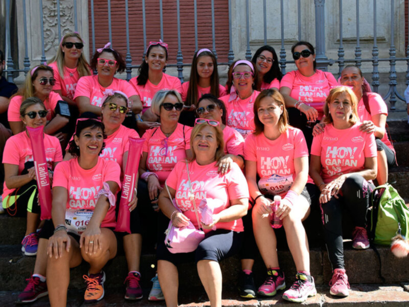 Una multitud de mujeres en camisetas rosas y gorros de lazo, posando en las escaleras de un edificio histórico, en una actividad de la 'Carrera de la Mujer'.