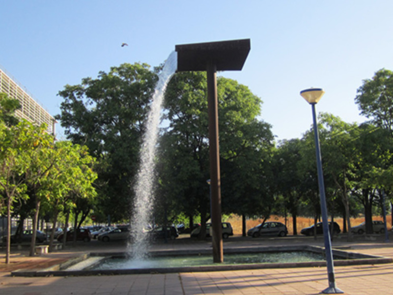 Fountain in a park with trees and a lamppost, surrounded by parked cars and a building in the background.
