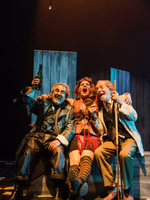 Three actors on stage, one holding a bottle, another with a cane, and one in a red dress, all appearing to be in a theatrical performance.