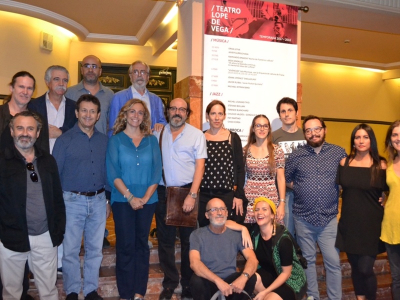 Grupo de personas posando para una foto en el Teatro Lope de Vega, con carteles de eventos culturales en el fondo.