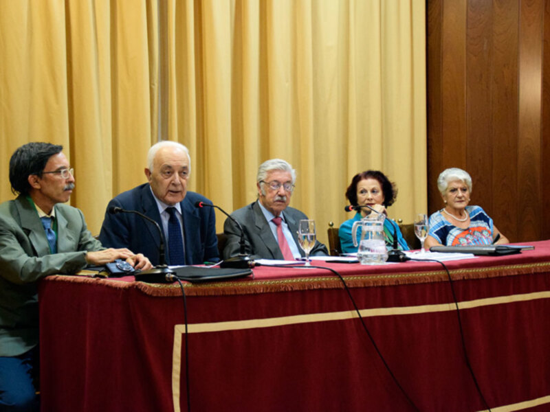 Conferencia de expertos sentados en una mesa con micrófonos, en un ambiente formal con cortinas doradas y un fondo de madera.
