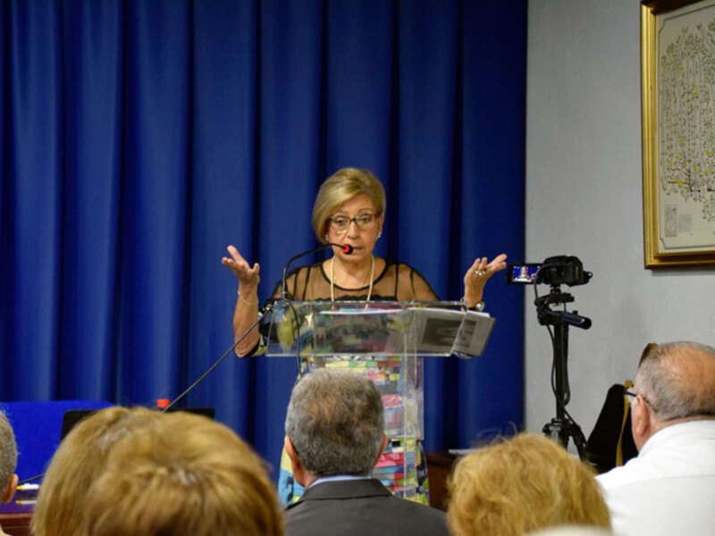 Mujer dando una conferencia en un escenario con cortina azul y público en primer plano.