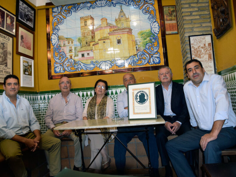 Grupo de personas sentadas en una mesa con un marco de fotos en el fondo, decoración de azulejos y carteles en la pared. El ambiente parece ser un lugar social o de reunión.